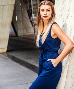 Street Fashion in New York. Young Eastern European American Woman with long brown hair, wearing blue sleeveless fashionable jumpsuit, standing against column outside office building, taking work break.