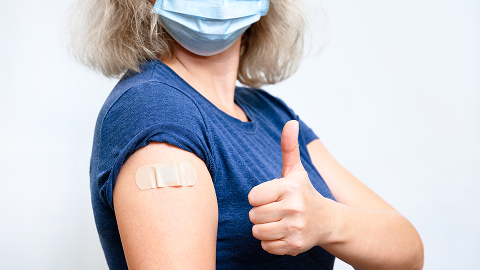 Frau mit blauem Oberteil und Pflaster von der Impfung mit Daumen hoch-Geste