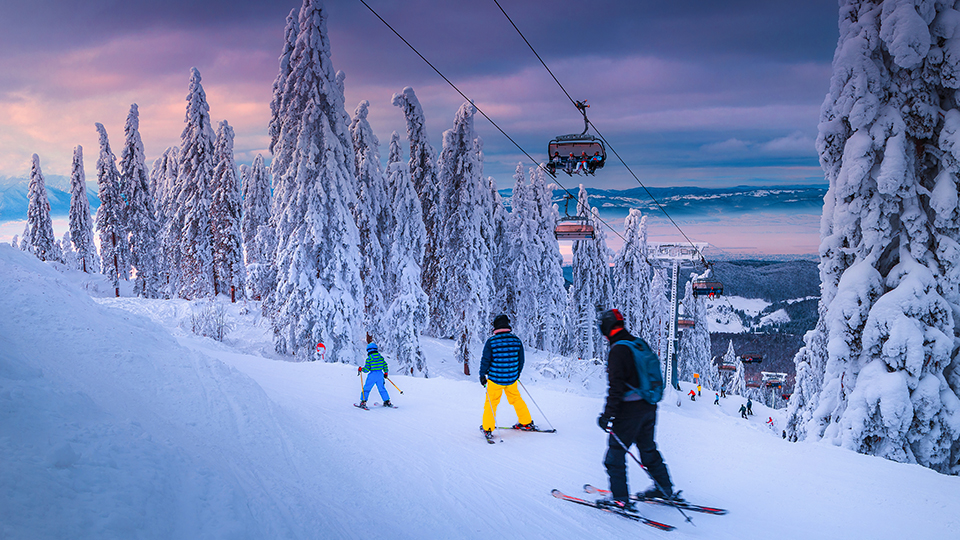 Schneelandschaft mit einer Gondel und drei Skifahrern. Der Himmel ist schon leicht lila eingefärbt.