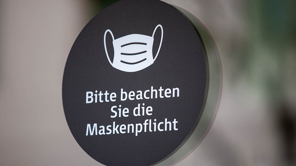 Abbilidung eines runden schwarzen Schildes mit weißer Aufschrift: Bitte beachten Sie die Maskenpflicht.