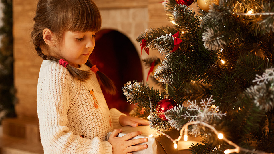 Kleines Mädchen mit weißem Pulli hält ein Geschenk in der Hand und betrachtet es andächtig. Im Hintergrund sieht man den Teil eines Christbaums.