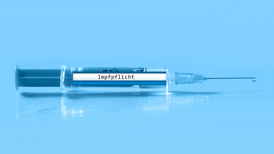 Dunkelblaue liegende Spritze vor hellblauem Hintergrund, auf der "Impfpflicht" steht