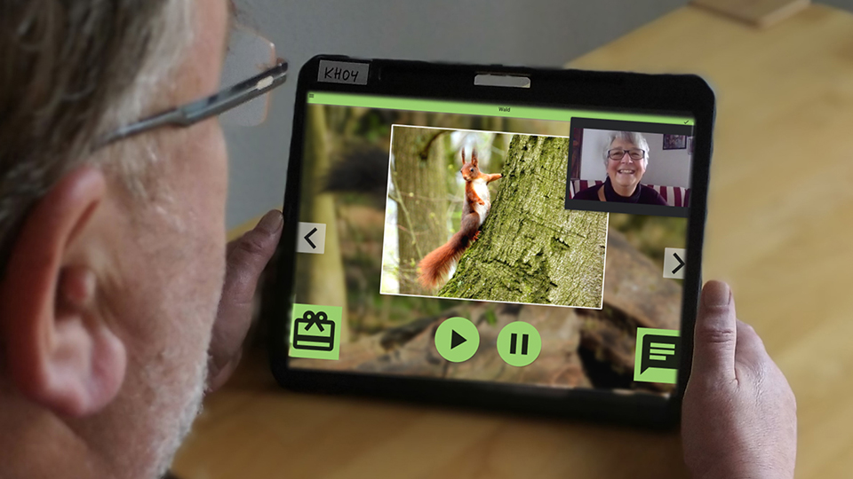 Schulterblick über einen älteren Herrn mit Brille, der auf ein Tablet schaut, wo er mit einer anderen Dame virtuell kommuniziert.