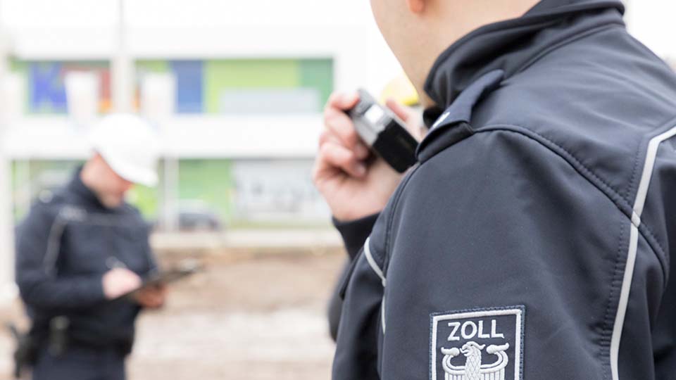 Zwei Männer vom Hauptzollamt. Einer ist schräg von hinten zu sehen und der Aufnäher seines Zoll-Wappens ist im Vordergrund zu sehen.