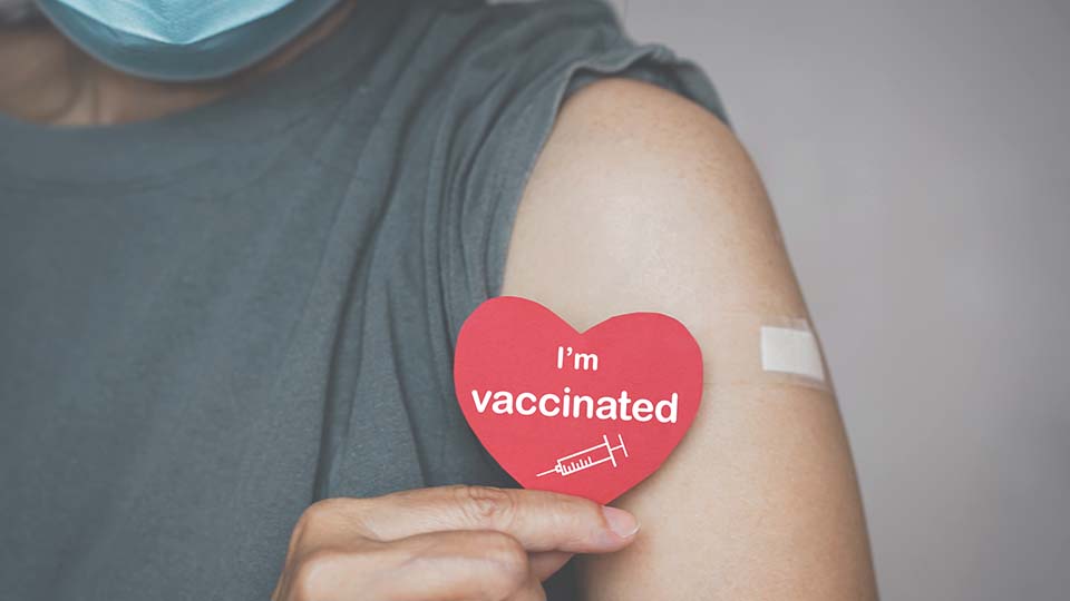 Person mit Maske und Pflaster auf dem Oberarm nach einer Corona-Impfung. Die Person hält ein Herz vor ihrem Arm, auf dem steht: "I am vaccinated."