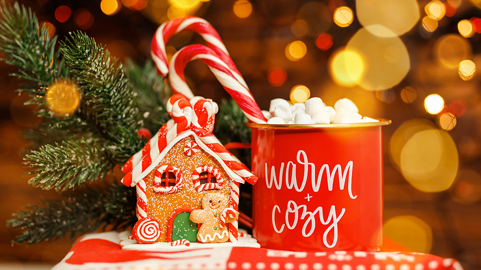 Rote Tasse mit Zuckerstangen darin und der Aufschrift: "Cozy". Daneben ein Lebkuchenhaus vor einem warmem rot-gelbem Hintergrund.
