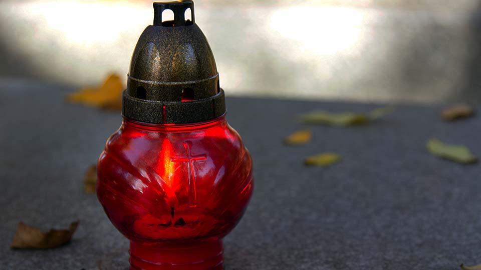 Rote Grabkerze auf dem Boden und dahinter Laubblätter am Boden.