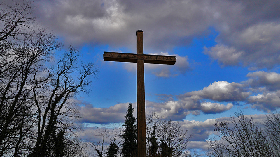 Großes Kreuz aus Holz vor einem blauen Himmel mit Wolken und kahlen Bäumen im Hintergrund.