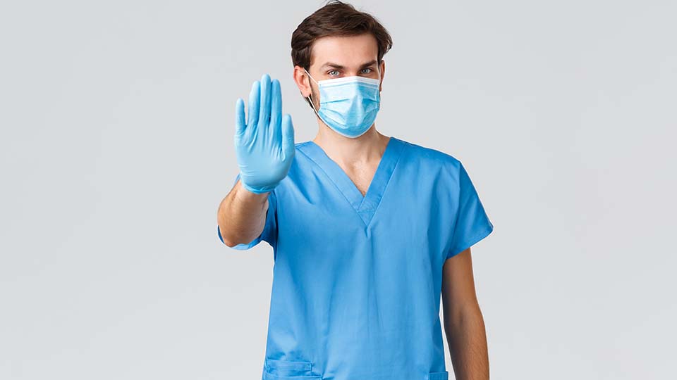 Arzt in blauem Oberteil und mit Maske hält die Hand als "Stopp-Zeichen" nach oben.