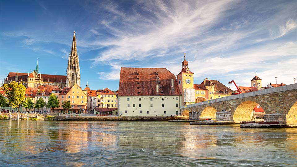 Bild von Regensburg von der Donau aus. Es ist im Vordergrund die Donau zu sehen und im Hintergrund die Stadtkulisse mit den beiden Domspitzen.