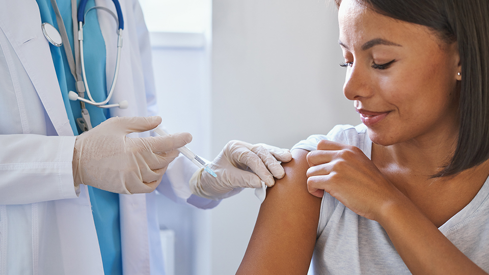 Eine dunkelhaarige Frau mit grauem Oberteil wurde gerade geimpft. Von der Ärztin ist nur der Kittel und die weißen Handschuhe zu sehen.