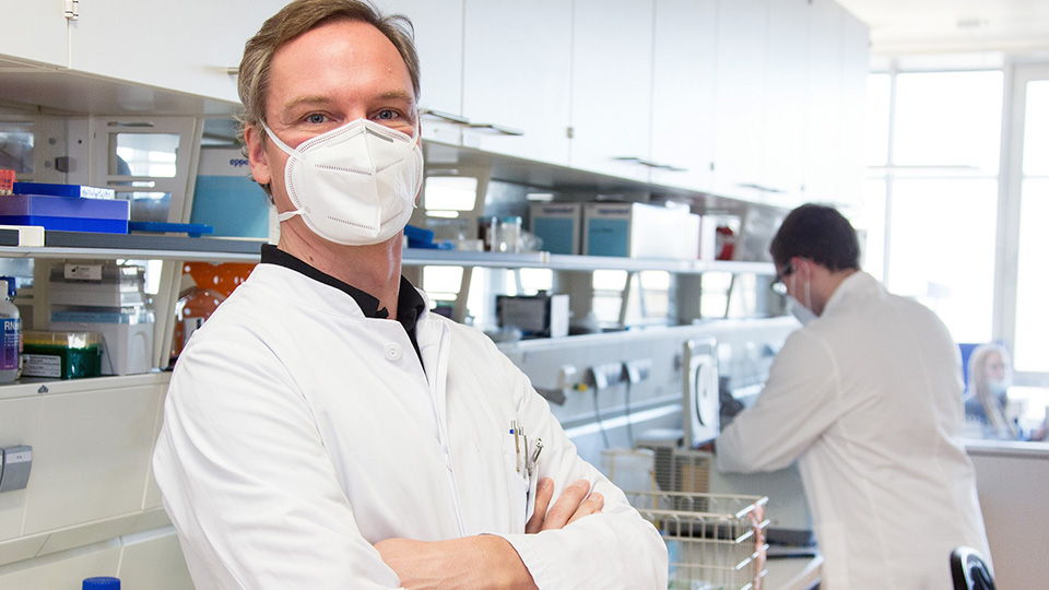 Professor Dr. Hendrik Poeck vom Universitätsklinikum Regensburg mit Maske. Dahinter sieht man einen Mitarbeiter im Labor.