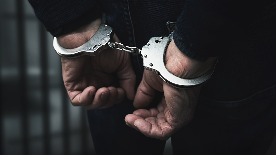 Zwei Hände in Handschellen in dem Rücken. Im Hintergrund Gefängnisgitter.