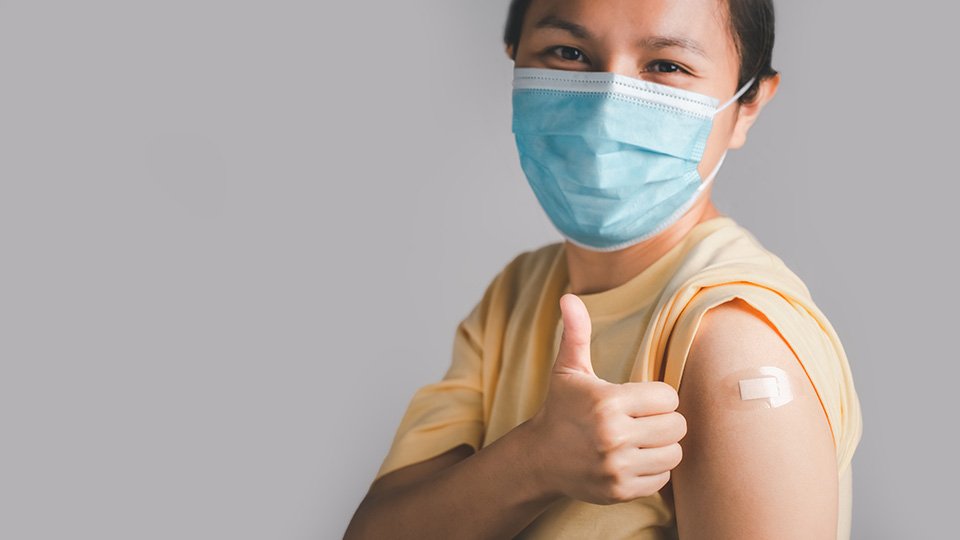 Dunkelhaarige Frau mit gelbem Oberteil und Maske, die ihr Pflaster am Oberarm nach der Corona-Impfung zeigt und das "Daumen hoch"-Zeichen gibt.