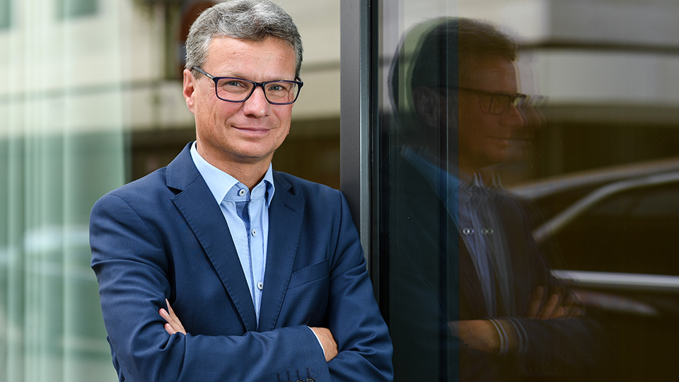 Staatsminister für Wissenschaft und Kunst, Bernd Sibler, wie er mit verschränkten Armen lächelnd an einer Scheibe lehnt.