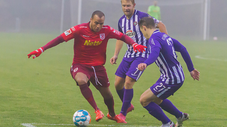 Jann George während er gerade in seinem roten Fußball-Trikot des SSV Jahn Regensburg den Ball spielt und von zwei gegnerischen Spielern angegriffen wird.