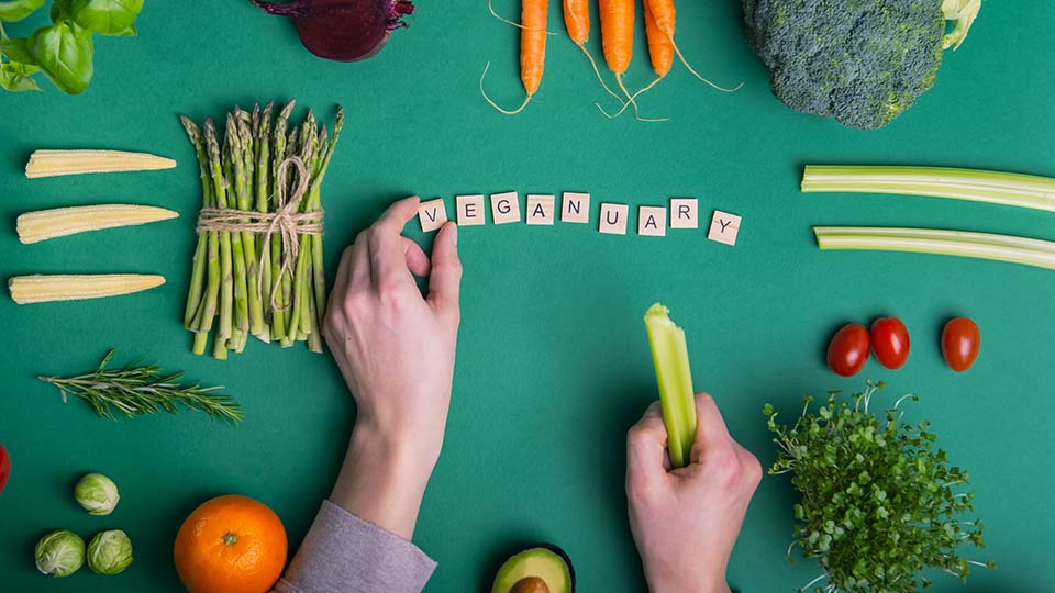 Zwei Hände, die das Wort "veganuary" mit Holzsteinen in die Mitte legen. Um das Wort herum liegen verschieden Obst- und Gemüsesorten, wie etwa Brokkoli, Rote Beete oder eine Avocado..