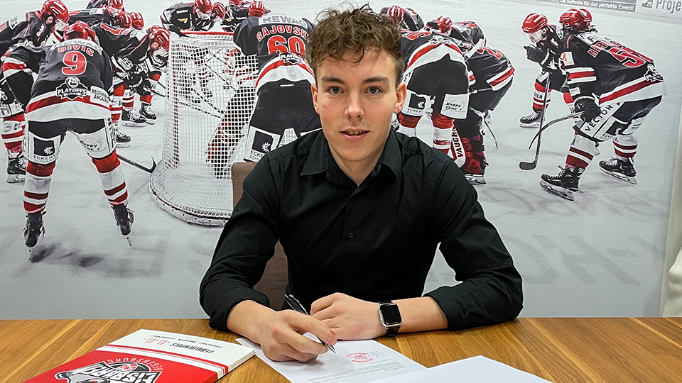 Der Stürmer der Eisbären Regensburg, Constantin Ontl, sitzt an einem Tisch und unterzeichnet seine Vertragsverlängerung. Im Hintergrund ist das Bild von einem Eishockeyspiel.