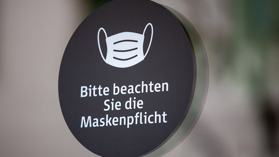 Schwarzes rundes Schild, auf dem eine weiße Maske abgebildet ist mit der Unterschrift: "Bitte beachten Sie die Maskenpflicht"
