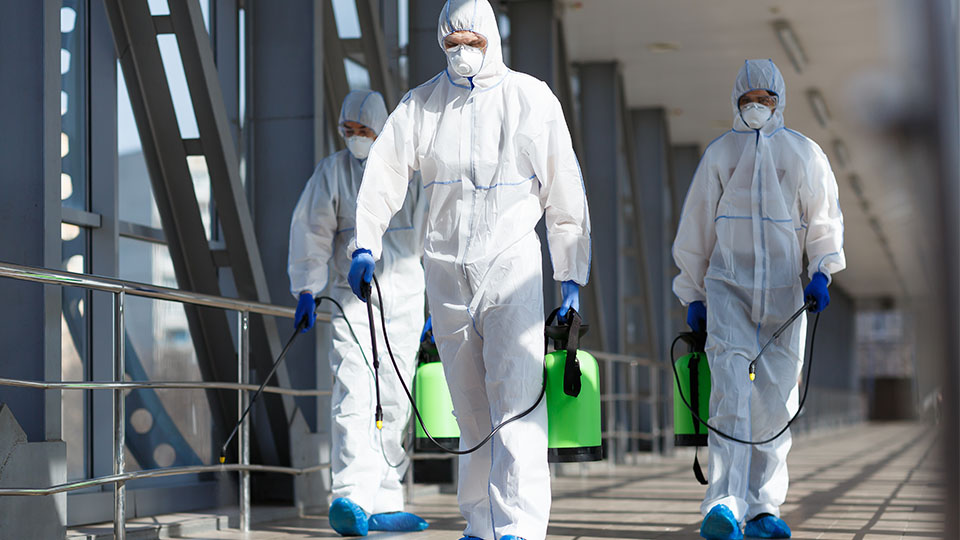 Drei Männer gehen in weißen Schutzanzügen und Masken sowie weiterer Ausrüstung zur Dekontamination einen Gang entlang.
