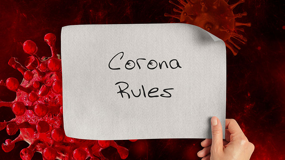 Rote Corona-Viren im Hintergrund und im Vordergrund wird in Zettel hochgehalte, auf dem steht: "Corona Rules", also Corona Regeln.