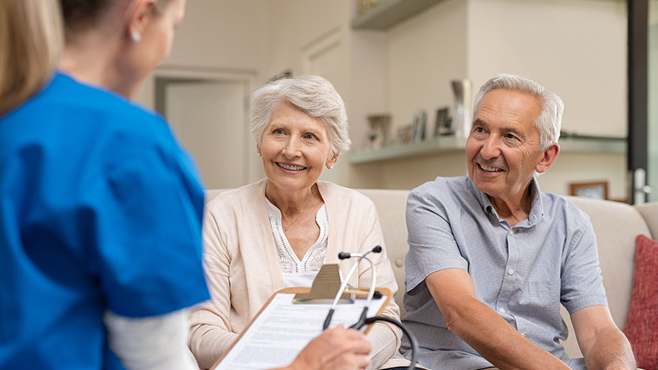 Pflegerin, die mit dem Rücken zur Kamera sitzt und etwas notiert. Im Fokus ist ein älteres Pärchen das lächelnd zu der Frau blickt.