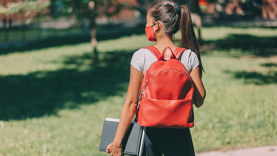Studentin am Campus der Hochschule von hinten mit Maske und ihren Unterlagen in der Hand.