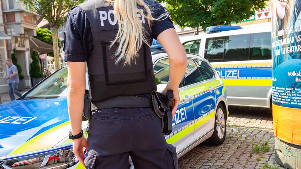 Blonde Polizistin von hinten mit der Hand an ihrer Pistole. Im Hintergrund stehen mehrere Streifenwagen der Polizei.
