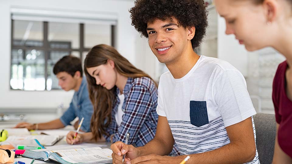 Junger Mann mit einem weißen T-Shirt, der gerade neben seinen Schulkameraden oder Kommilitonen sitzt. Er hat einen Stift in der Hand und lächelt.
