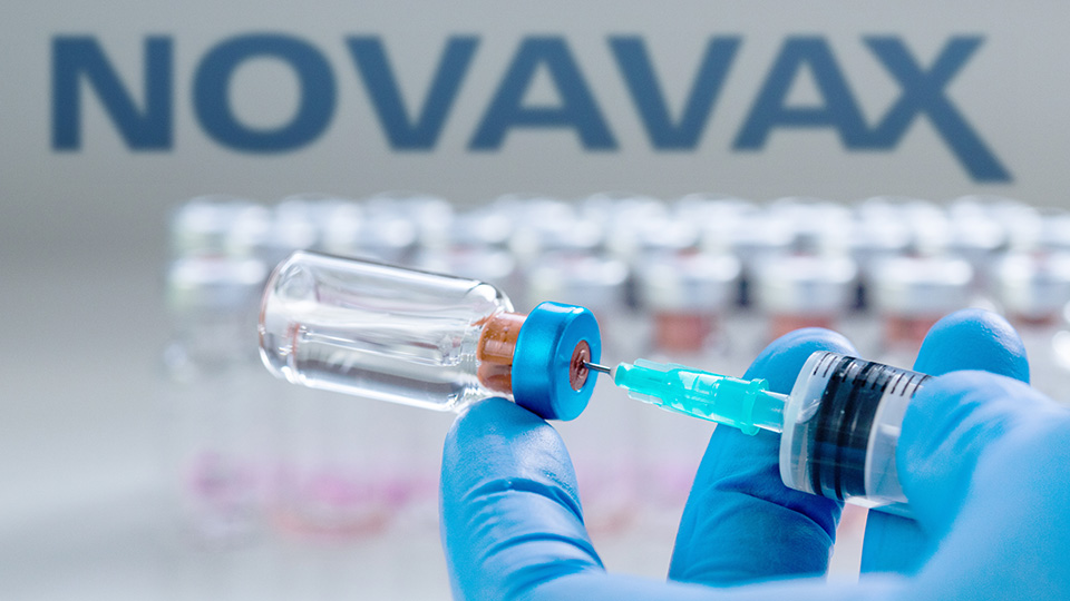 Eine Hand mit blauem Gummihandschuh zieht gerade eine Impfung mit Novavax auf. Im Hintergrund steht der Schriftzug "NOVAVAX".