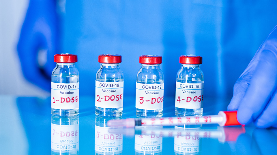Vier Dosen mit Corona-Impfstoff nebeneinander und davor liegend eine Spritze
