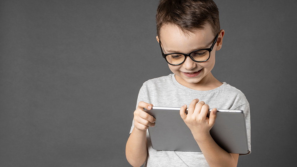 Umgang mit Medien: Dunkelhaariger Junge mit Brille hält Tablet in der Hand und lacht.