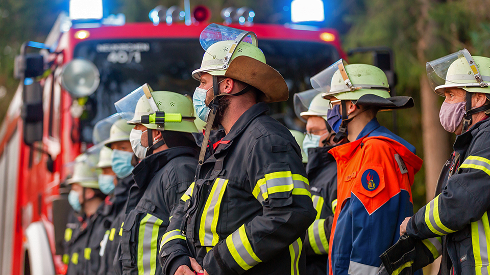 Mehrere Feuerwehrmänner in ihrer Feuerwehrausrüstung und mit Masken. Dahinter steht ein Feuerwehrfahrzeug mit Blaulicht.