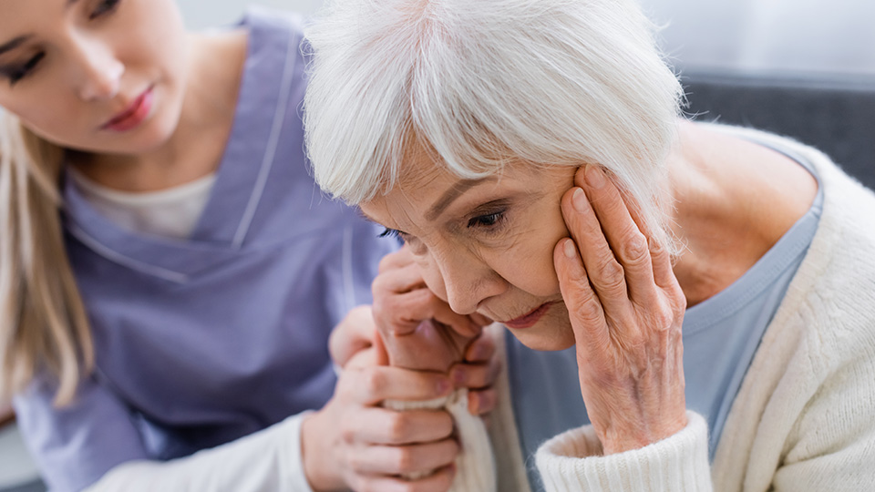 Angehörige junge Frau, die die Hand einer älteren Damen hält, die sich mit beiden Händen den Kopf hält, was ihre Demenz-Erkrankung symbolisiert.