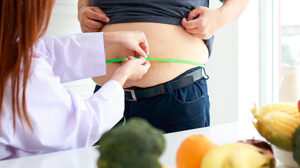 Ärztin misst Bauchumfang eines Mannes mit starkem Übergewicht. Im Vordergrund liegt Gemüse auf dem Tisch.