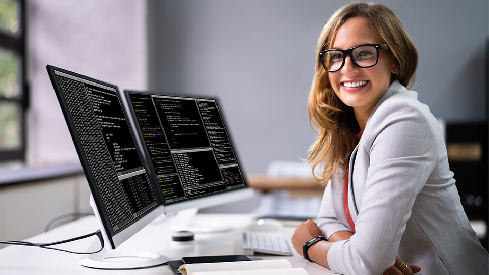 Blonde Programmiererin mit Brille, die gerade vor ihren zwei Bildschirmen sitzt und lächelt.