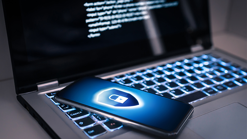 Aufgeklappter Laptop, auf dem ein Virencode sichtbar ist und darauf leigend ein Handy, auf dem als Schutz vor Cyberangriffen eine Sperre aktiviert ist.