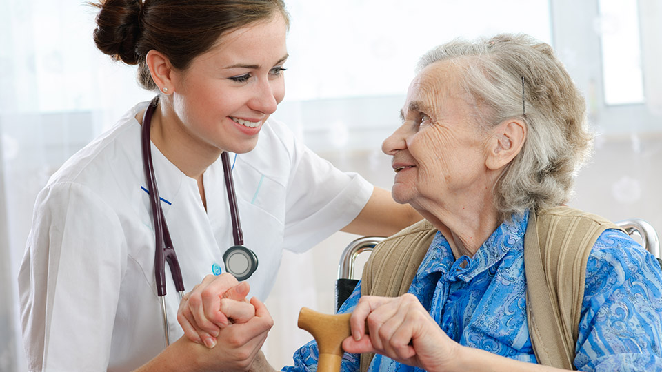 Ärztin, die einer älteren Dame in einem Pflegeheim die Hand hält und sie anlächelt.