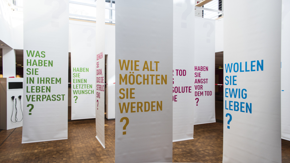 Vernissage - Wollen Sie ewig leben? - "Dialog mit dem Ende". Fragen wie "Was haben Sie in ihrem Leben verpasst" sind aufgehängt.