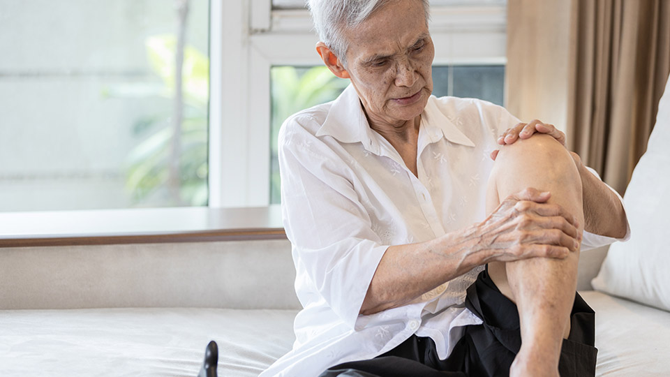 Ältere Frau, die unter Rheuma leidet und daher und Schmerzen im Knie. Sie umgreift mit beiden Händen ihr Knie.