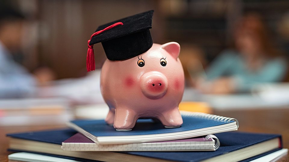 Finanzierung des Studiums: Sparschwein mit Absolventenhut auf einem Bücherstapel.