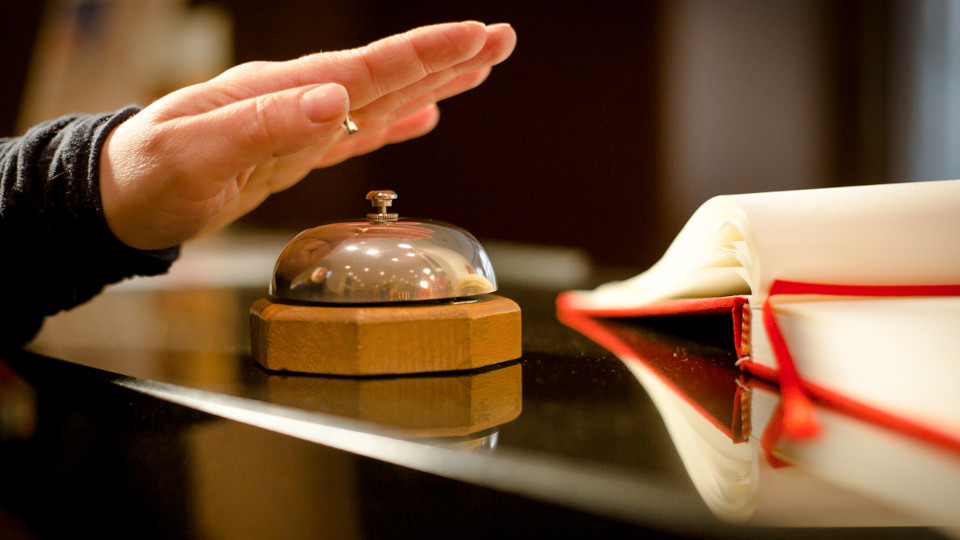 Fehlendes Personal in Hotels: Hand, die gerade auf eine Empfangsglocke tippt, daneben ein Reservierungsbuch.
