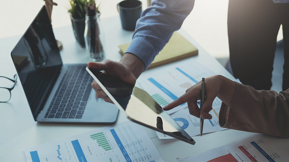 Lieferantenmanagement: Zwei Personen am Schreibtsich, die über Statistiken, Laptop und Ipad sitzen und diskutieren.