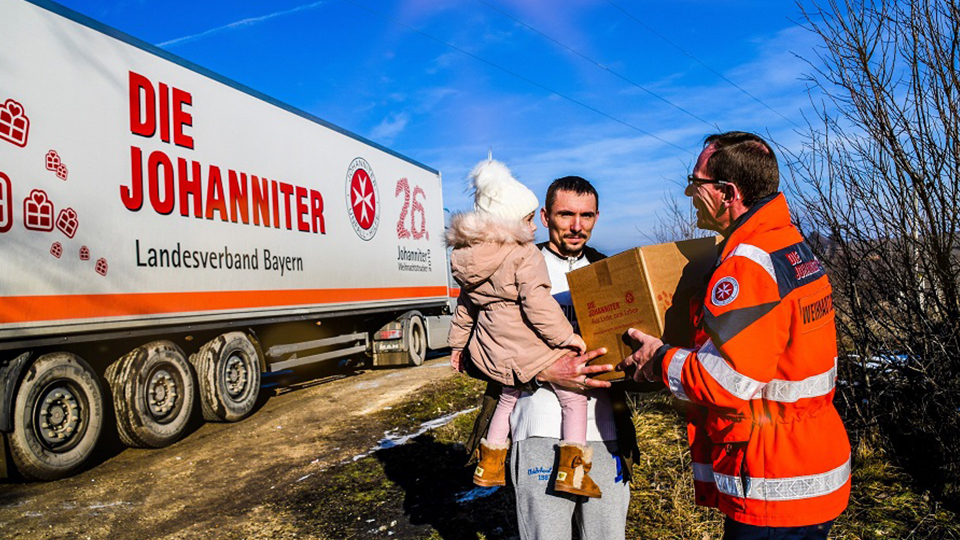 Johanniter Weihnachtstrucker: Ein Freiwilliger Helfer der Johanniter übergibt einem Mann mit Kind auf dem Arm sein Paket.