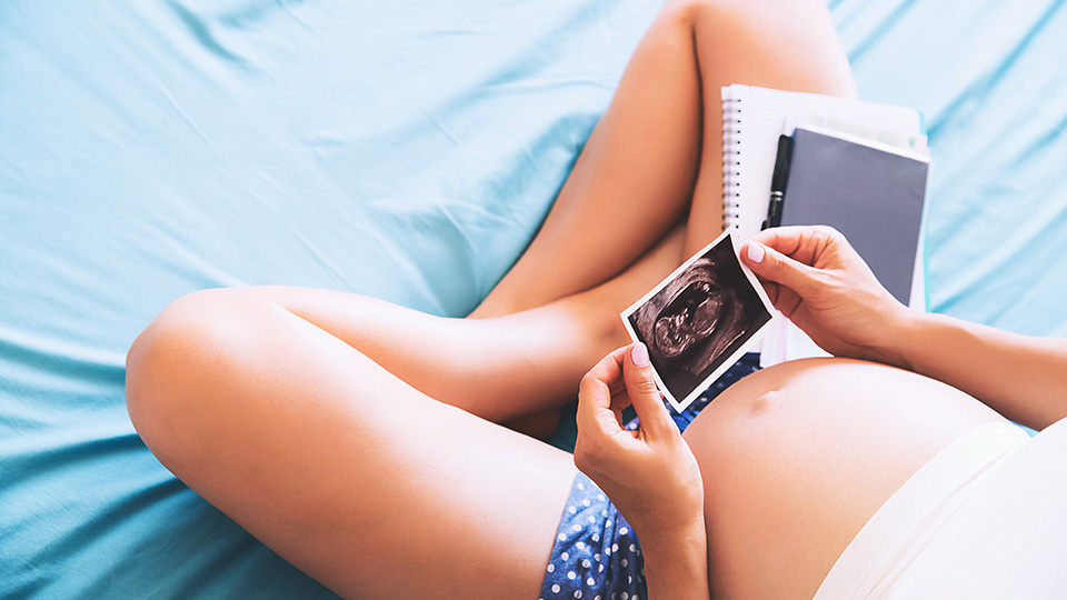 Mythen rund um den Kinderwunsch: Schwangere Frau mit Ultraschallbild sitzt auf dem Bett
