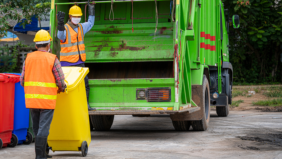 Zwei Männer der Müllabfuhr, die die Gelbe Tonne zum Entleeren zum Müllfahrzeug schieben.