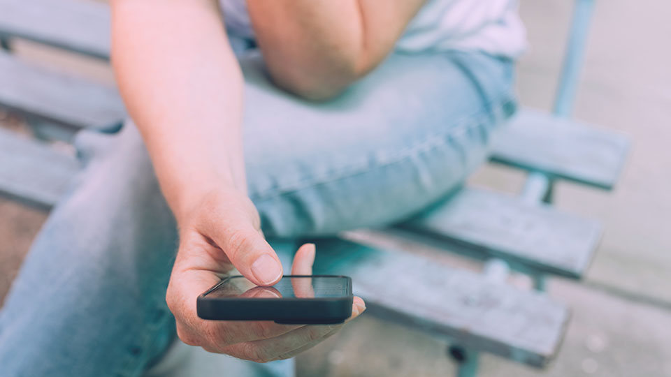 FOMO: Junge Frau, die alleine auf einer Bank sitzt und auf ihr Handy schaut