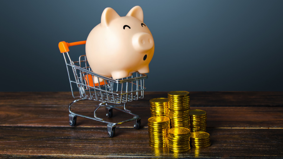 Sparschwein, das in einem Einkaufswagen auf Stapel aus Münzen zurollt.