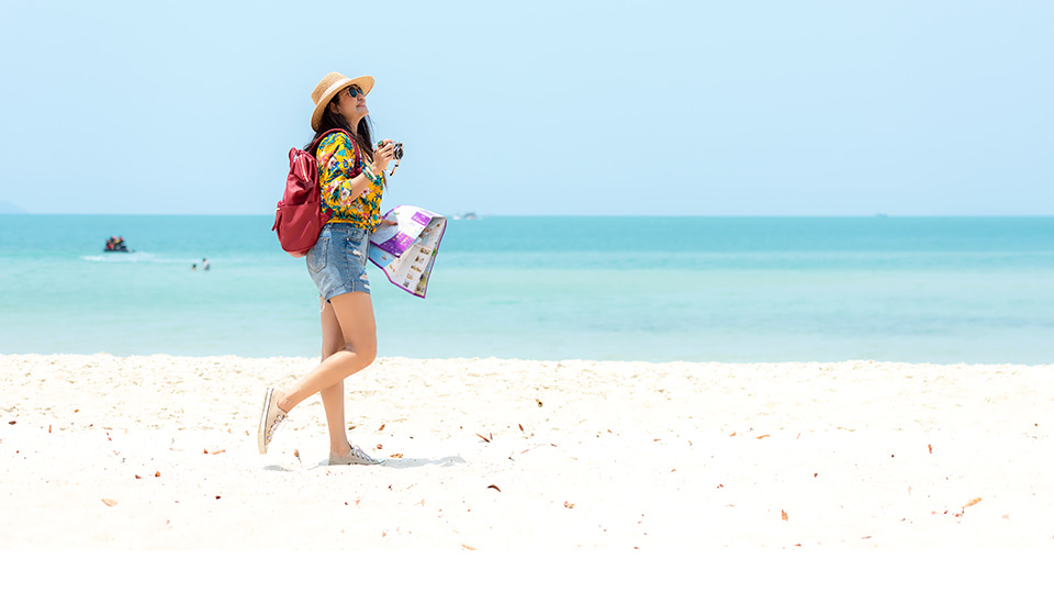 Sommerurlaub: Eine Frau geht lächelnd mit einem Sonnenhut auf dem Kopf am Strand entlang, im Hintergrund Meer.