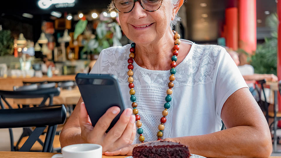 Seniorin macht einen Videochat über ihr Handy und sitzt lächeln mit einem Stück Kuchen in einem Café